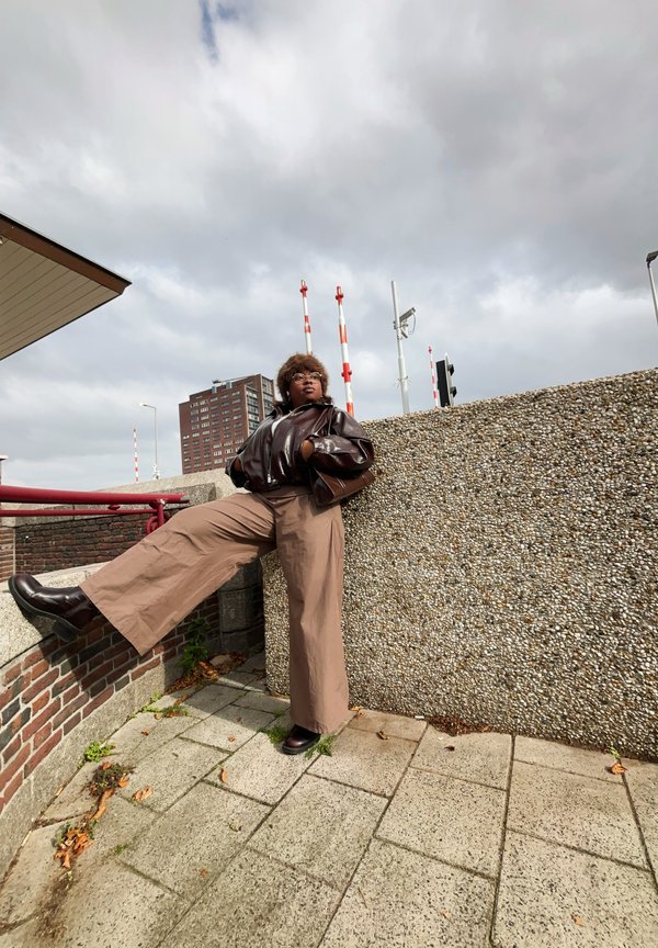 Barna fényes dzseki, bézs széles szárú nadrág, sötét csizmák; egy strukturált betonfal előtt pózol piros és fehér csíkos oszlopokkal a háttérben.