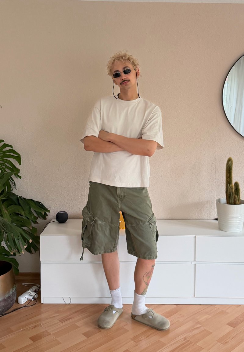 Personne avec des cheveux blonds bouclés portant des lunettes de soleil, un t-shirt blanc, un short cargo vert, des chaussettes blanches et des chaussons, debout les bras croisés à l'intérieur.