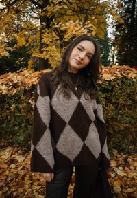 Jeune femme aux longs cheveux bruns portant un pull à losanges marron et beige, debout à l'extérieur parmi les feuilles d'automne et les arbres.