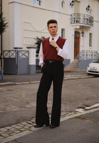 Muž v červené vestě přes bílou košili upravující si kravatu, oblečený v černých kalhotách a botách, stojící na ulici poblíž obytných budov.