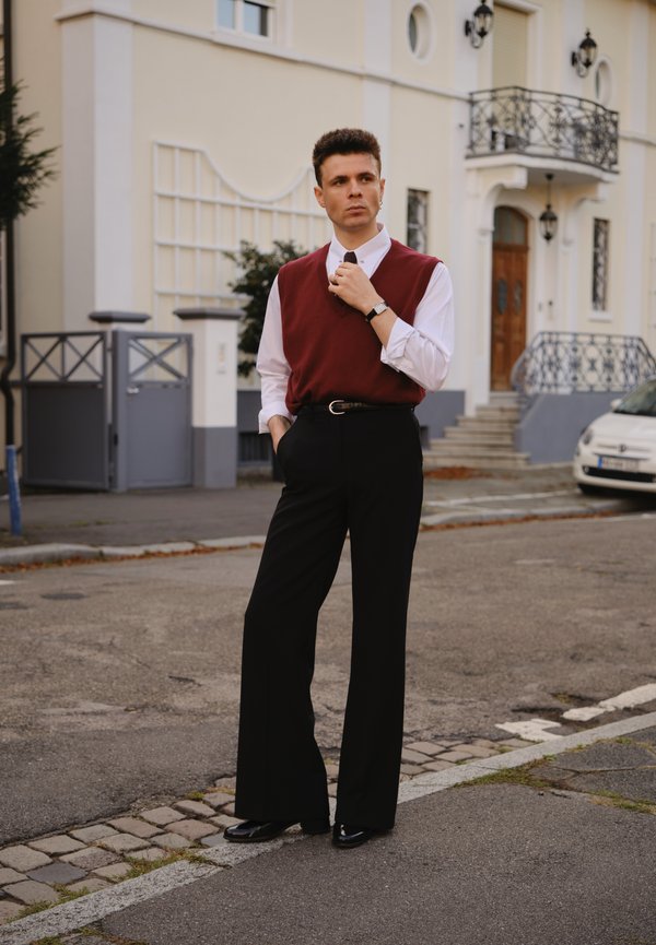 Hombre con chaleco rojo sobre camisa blanca ajustándose la corbata, vistiendo pantalones y zapatos negros, de pie en una calle cerca de edificios residenciales.