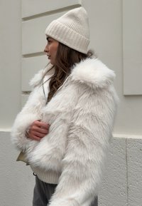 Λευκό ανάγλυφο μπουφάν από ψεύτικη γούνα με κουκούλα, ριμπ μανσέτες και υφή εξωτερικά. Φορεμένο με ένα ανοιχτό κρεμ πλεκτό σκουφί. Εμφάνιση από το πλάι.