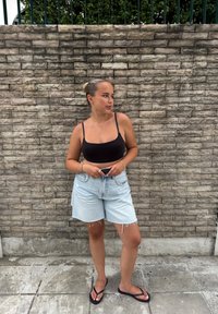 Musta lyhyt hihaton toppi ja vaaleansiniset farkkushortsit, joiden reunat ovat rispaantuneet, yhdistettynä mustiin varvassandaaleihin, takanaan teksturoidulla kiviseinällä.