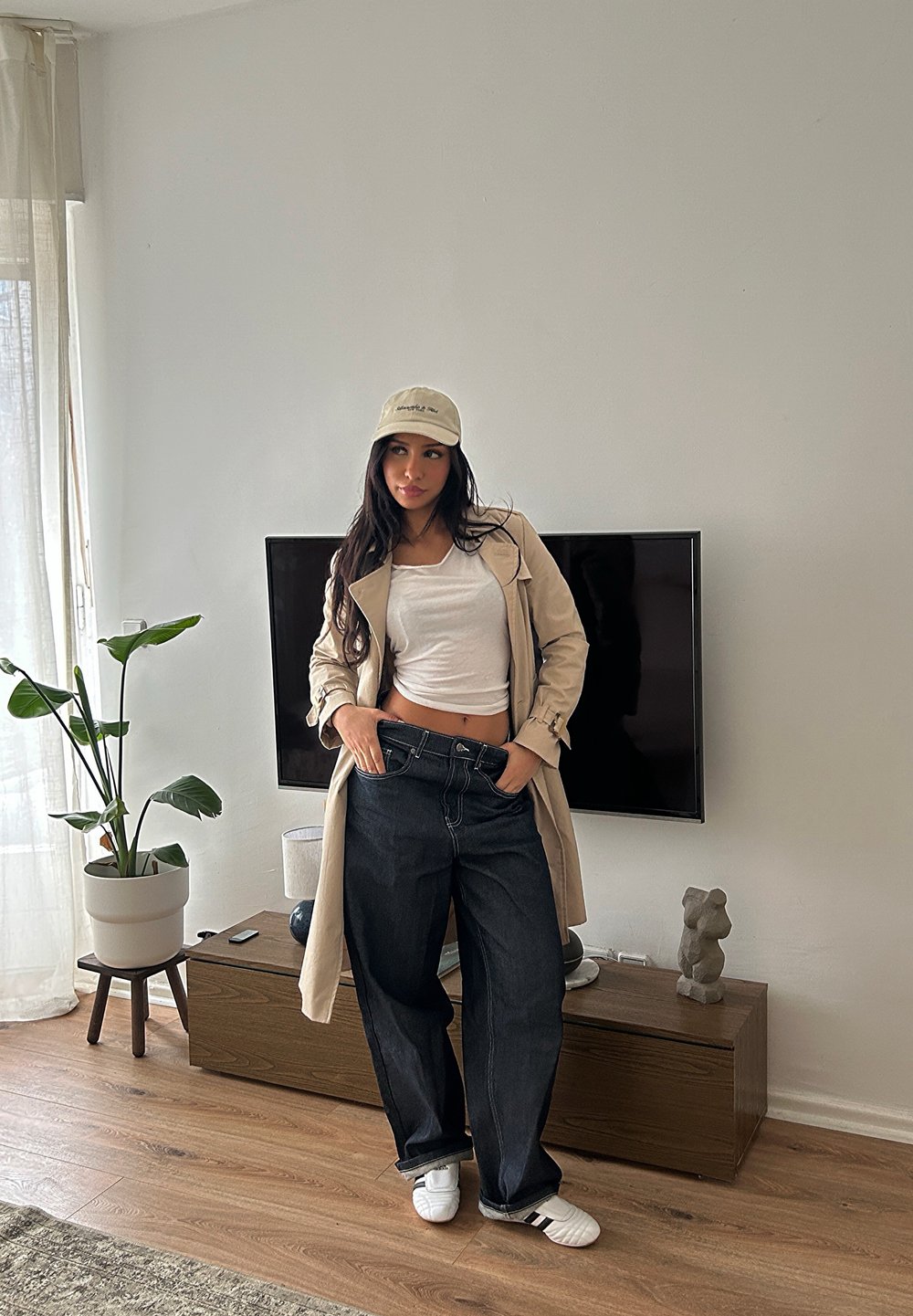 Trench beige, top court blanc et jean en denim foncé. Debout sur un sol en bois, avec une plante en pot et une télévision en arrière-plan.