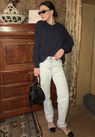 Mujer con cárdigan negro y jeans blancos posa en interiores sosteniendo un bolso negro, lleva gafas de sol y zapatos planos bicolores beige y negro.