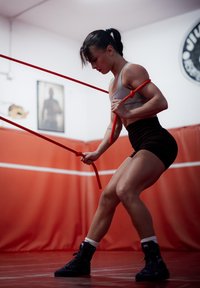 Vaja z rdečimi upornimi trakovi. Oseba nosi sivo majico brez rokavov in črne kratke hlače v telovadnici z rdečimi stenami.