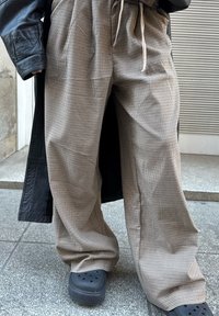 Partie inférieure du corps portant un pantalon large à carreaux beige, des chaussures Crocs noires et une veste en cuir noire partiellement visible.