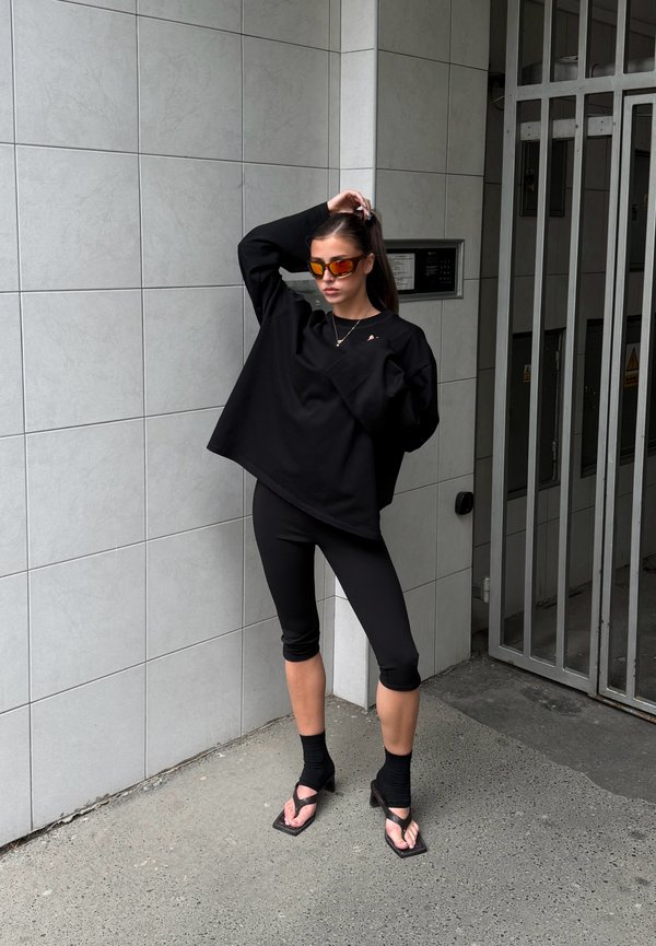 Frau trägt schwarzen übergrossen Top, schwarze Capri-Leggings, schwarze Sockensandalen und orange getönte Sonnenbrille, steht vor einer gefliesten Wand neben einem Metalltor.