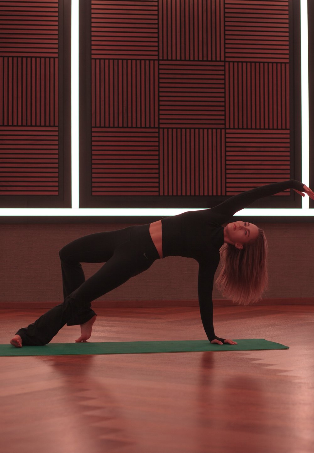 Femeie îmbrăcată în echipament de sport negru, executând o poziție de yoga laterală pe o saltea verde, într-o cameră cu podea de lemn și panouri acustice roșii.