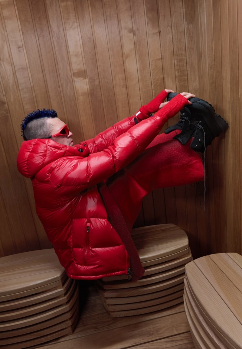 Veste matelassée rouge avec capuche, assortie à une tenue rouge et des bottes à plateforme noires. Lunettes de soleil rouges et cheveux bleus complètent le look. Intérieur en bois.