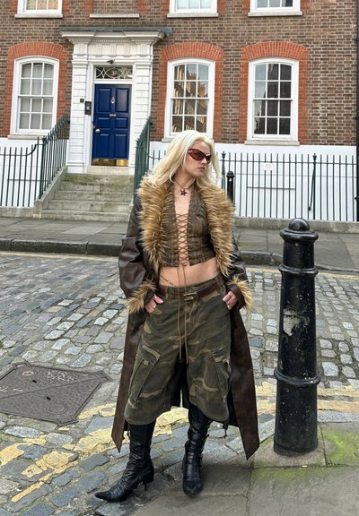 Abrigo de cuero marrón con cuello de piel sintética, pantalones cortos de camuflaje verde, y botas negras hasta la rodilla, complementado con gafas de sol y un colgante de estrella.