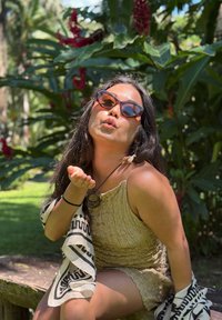 Femme portant des lunettes de soleil et une robe beige souffle un baiser en étant assise sur un banc en bois entouré de plantes tropicales vertes et de fleurs rouges.