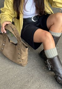 Brun suède håndveske med sølvnagler, kombinert med mørke shorts og brune ankelstøvler. Teksturert grå sokker fremhever stilen. Gul jakke synlig.