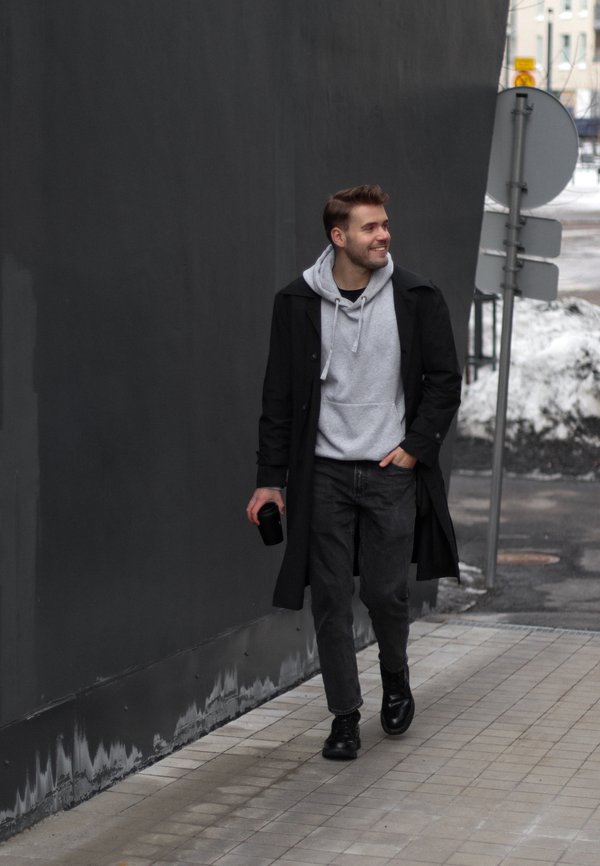 Schwarzer Mantel, grauer Kapuzenpullover, dunkle Jeans und schwarze Schuhe. Ein Mann hält einen schwarzen Becher und geht auf einem gepflasterten Weg neben einer dunklen Wand.