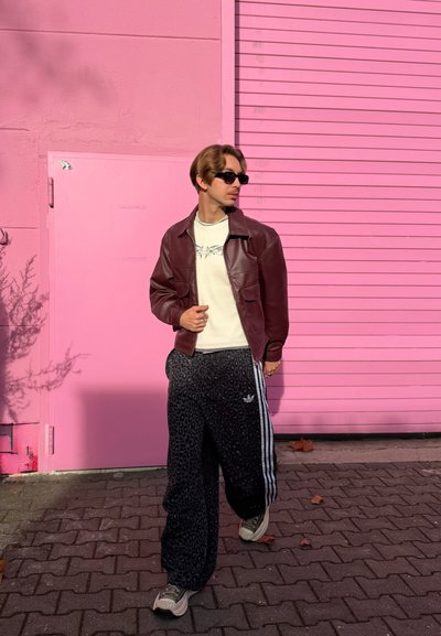 Chaqueta de cuero marrón sobre un suéter blanco, pantalones deportivos negros con patrón de leopardo y rayas blancas, y zapatillas grises contra un fondo rosa.