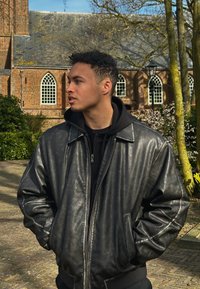Casaco bomber de couro preto com fecho, colarinho largo e punhos em ribana, usado sobre um hoodie preto, em frente a um fundo de parque verde.