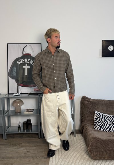 Hombre con pelo rubio que lleva una camisa marrón de cuadros y pantalones anchos blancos, de pie en el interior cerca de un sofá marrón y una estantería con decoraciones.