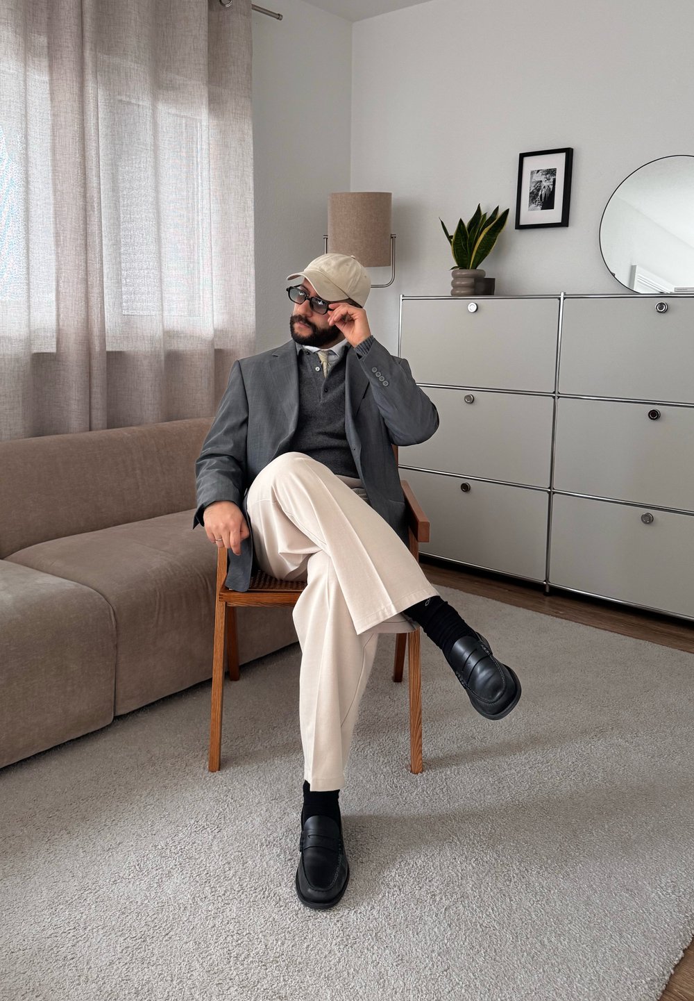 Homme portant un blazer, une casquette et des lunettes de soleil, assis sur une chaise en bois dans un salon moderne et minimaliste avec un canapé, un meuble de rangement et une plante.
