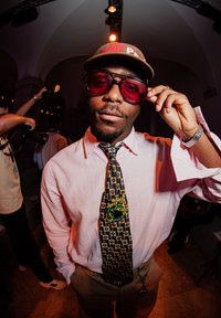 A man is wearing a light pink shirt, a patterned tie with gold accents, and a brown cap. He has dark sunglasses on and a silver watch. There is a green accent on his tie.