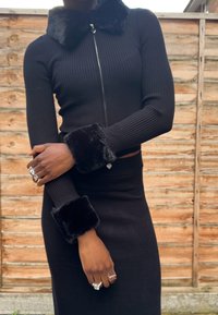 Čierny žebrovaný sveter na zips s umelým kožúškom na golieri a manžetách, skombinovaný s čiernou priliehavou sukňou. Hladká textúra a elegantný strih.
