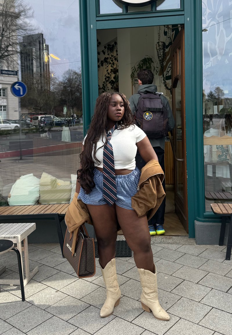 Femme aux longs cheveux portant une chemise courte blanche, une cravate, un short bleu, des bottes beige, tenant une veste et un sac, debout devant la porte d'un café.