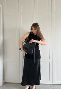 Musta hihaton toppi ja pitkä hame, molemmat sileästä kankaasta. Suuri musta olkalaukku pidetään sivussa, jossa on teksturoituja yksityiskohtia. Neutraali tausta.