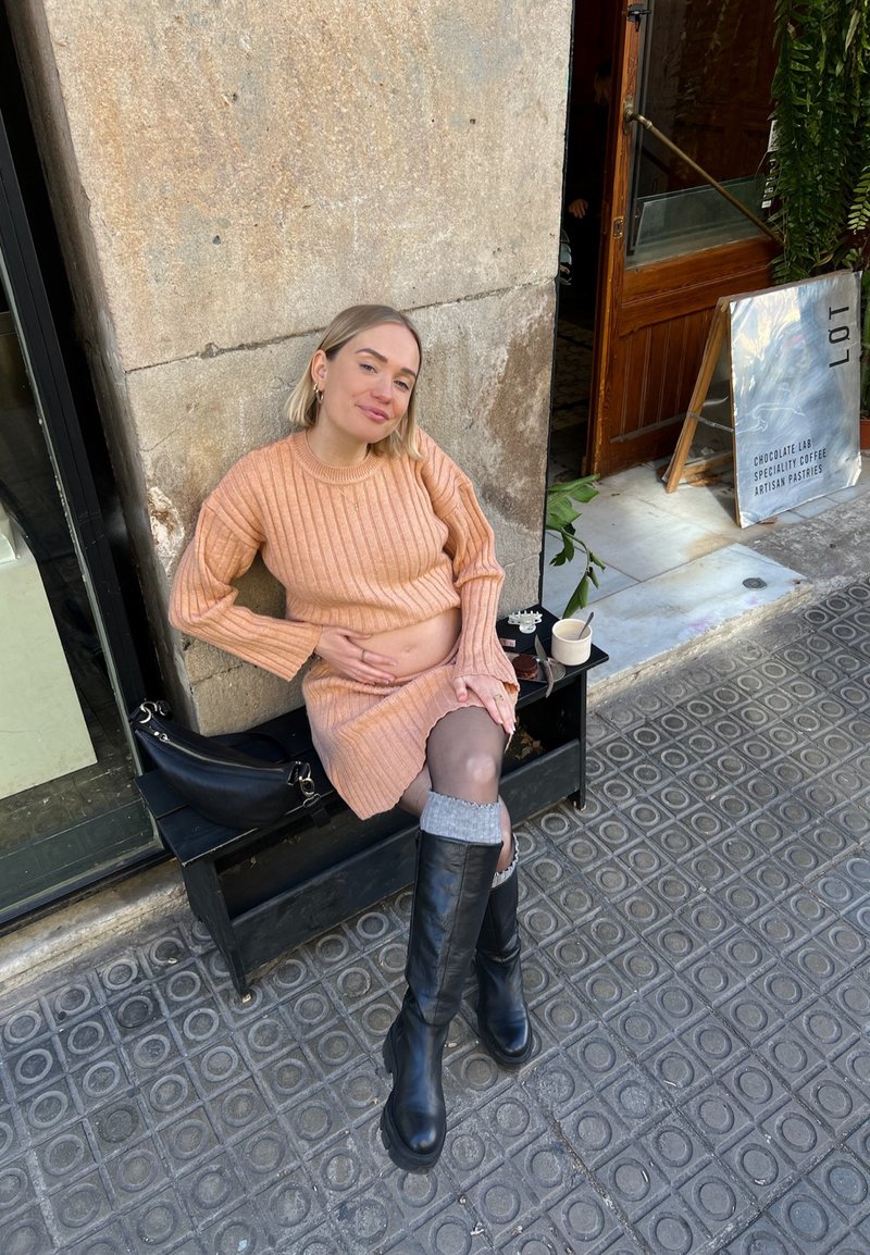 Πλεκτό ροζ φόρεμα με ριμπ ύφανση, μαύρες μπότες μέχρι το γόνατο και γκρι κάλτσες, καθισμένο σε μια πέτρινη τοιχοποιία με ένα φλιτζάνι καφέ.