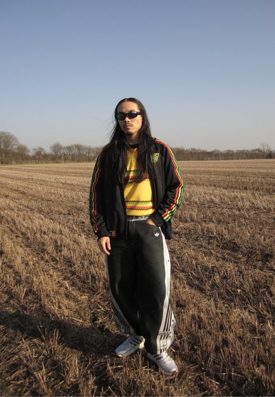Joven con cabello largo y gafas de sol de pie en un campo seco, vistiendo una chaqueta negra, suéter amarillo, pantalones negros de Adidas y zapatillas blancas.