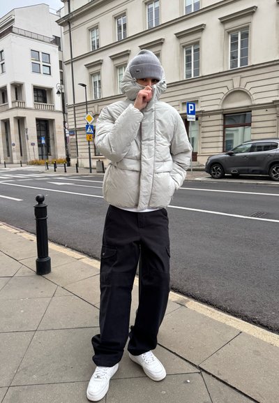 Chaqueta de plumas gris claro con cuello alto, llevada sobre una camisa blanca. Unos pantalones cargo negros y zapatillas blancas completan el atuendo. Entorno urbano.