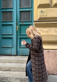 Dugi kaput od vune u smeškanom kariranom uzorku, u kombinaciji s plavim farmerkama. Osoba drži bijelu šolju pored istrošenih tirkiznih vrata.