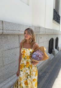Robe blanche avec des motifs de roses jaunes, corsage ajust�é et bretelles spaghetti. Tenant un bouquet de fleurs séchées violettes enveloppé dans du papier brun.