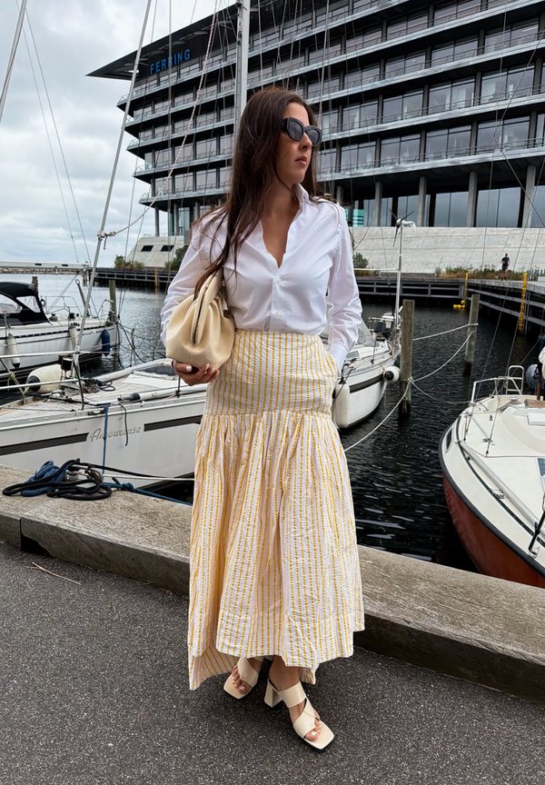 Woman in white blouse, yellow striped skirt, and sandals stands by marina with boats and modern building in the background.