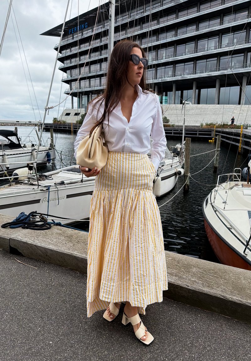 Woman in white blouse, yellow striped skirt, and sandals stands by marina with boats and modern building in the background.