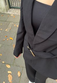 Černé sako na míru s velkým klopovým límcem a ozdobným zlatým špendlíkem v pase, nošené přes černý top a kalhoty.