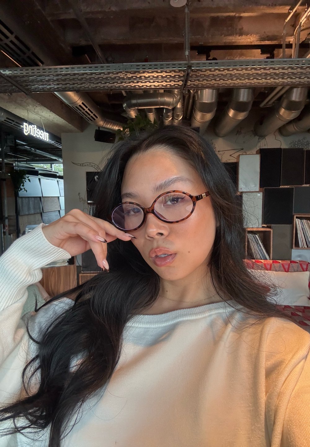 Gafas de carey con lentes redondos, suéter texturizado y cabello largo y oscuro; en interiores con tuberías de metal y fondos estampados.