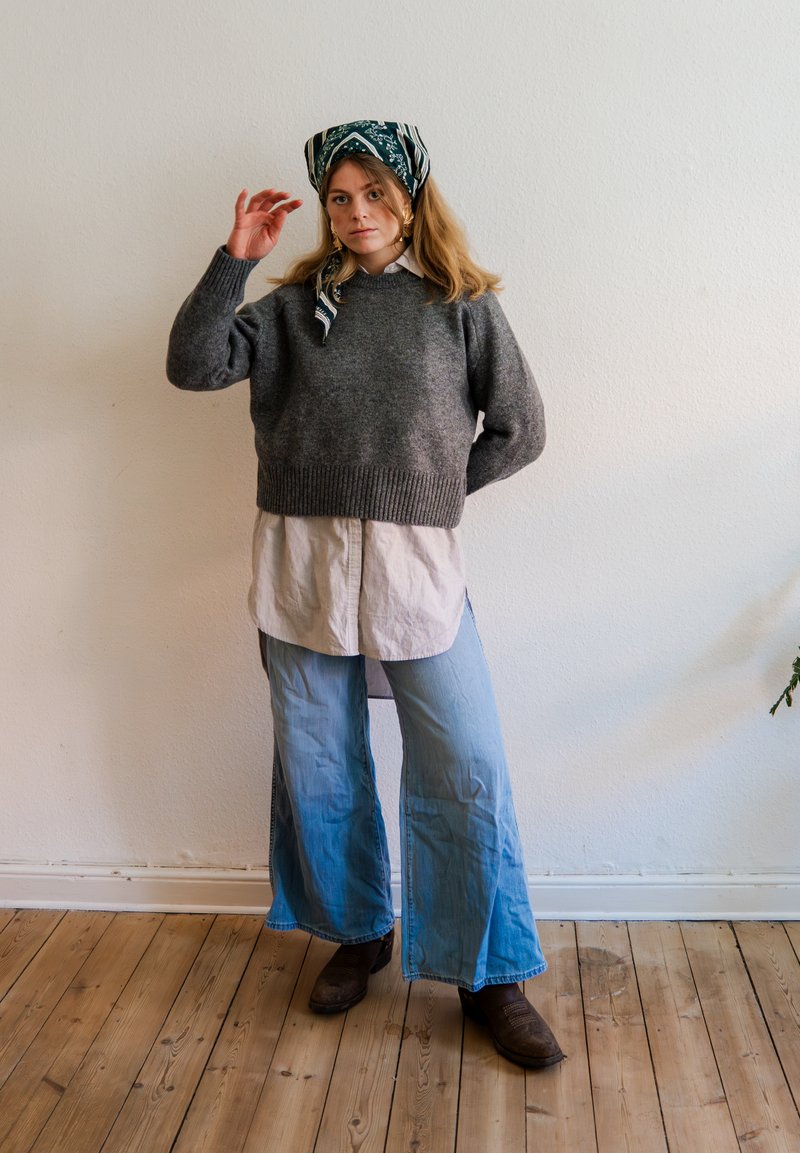 Femme portant un pull gris, une chemise blanche, un jean large bleu, des bottes marron et un foulard à motifs, debout sur un sol en bois contre un mur blanc.