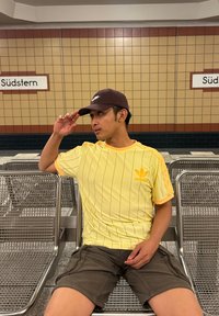 Žluté adidas tričko s pruhy a oranžovými akcenty, spárované s tmavými šortkami. Hnědá kšiltovka dotváří ležérní vzhled. V pozadí kovové sezení.