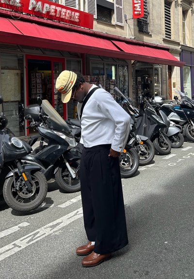 Camisa blanca de manga larga, pantalones anchos negros, zapatos marrones y una gorra beige. El fondo presenta scooters aparcados y una tienda con un toldo rojo.