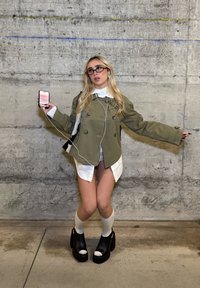 Oliivinvihreä ylikokoinen kaksirivinen takki, alla valkoinen kauluspaita, shortsit, mustat korokepohjaiset kengät, valkoiset polven korkeudelle ulottuvat sukat, kädessä puhelin.