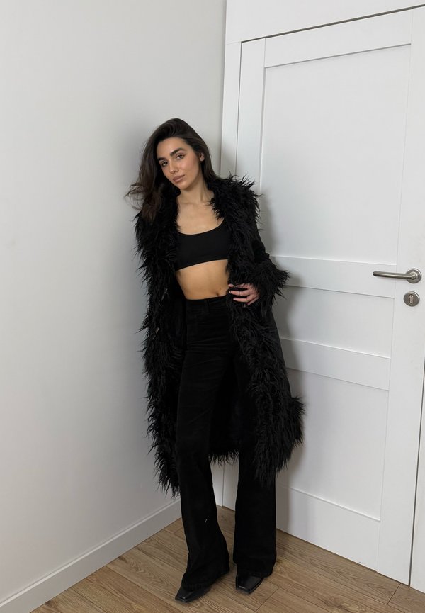 Femme debout à l'intérieur, contre un mur blanc et une porte, portant un haut court noir, un pantalon taille haute noir et un long manteau noir en fourrure.