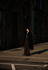 Personne portant un manteau foncé et un pantalon marron se tient sur un parking pavé, projetant une longue ombre, bâtiment en briques avec de grandes fenêtres en arrière-plan.