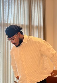 Un homme portant une casquette noire et des lunettes ajuste sa chemise blanche à fines rayures dans une pièce éclairée doucement avec des rideaux beiges et une chaise marron.