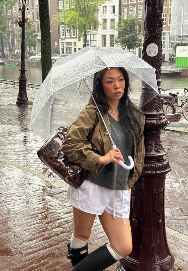 Femme en imperméable et bottes tenant un parapluie transparent dans une rue de ville mouillée près d'un canal et d'un lampadaire par jour de pluie.