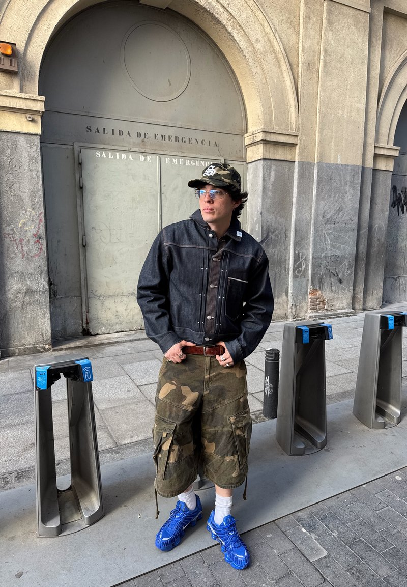 Ein junger Mann in Tarnshorts, einer dunklen Jacke, blauen Sneakers und einer Tarnkappe steht in der Nähe von Metall-Drehtoren und einer Notausgangstür.