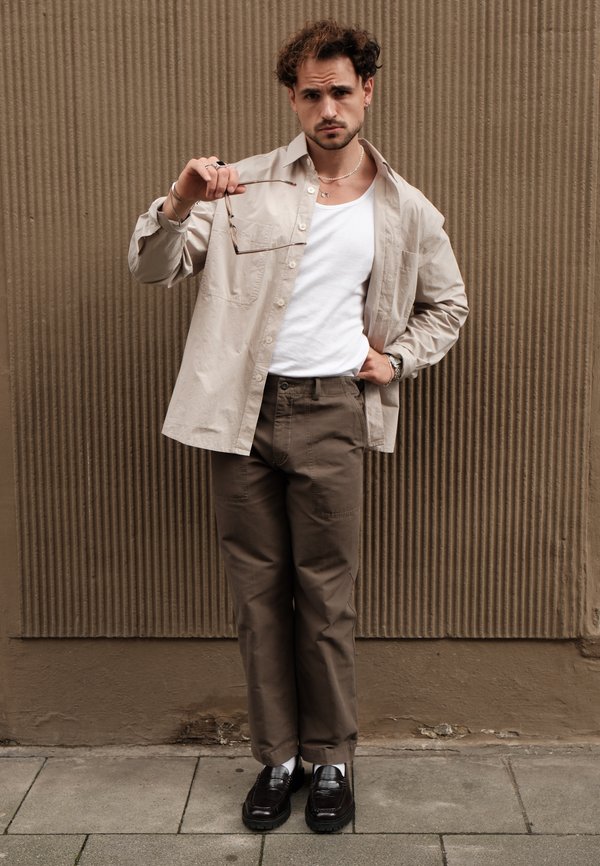 Camisa oversized bege, top branco, calças castanhas e mocassins pretos; segurando óculos com colares em camadas e brincos. Fundo texturizado.