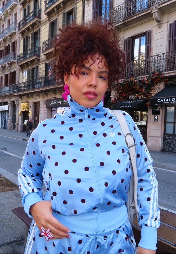 Vrouw met krullend haar draagt een blauw trainingspak met witte stippen en roze oorbellen, staand op een stadsstraat met gebouwen en winkels op de achtergrond.