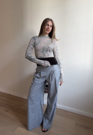 Mujer con blusa de manga larga con estampado floral, pantalones grises de pierna ancha y cintura alta, y sandalias negras, de pie en una habitación con mobiliario minimalista.