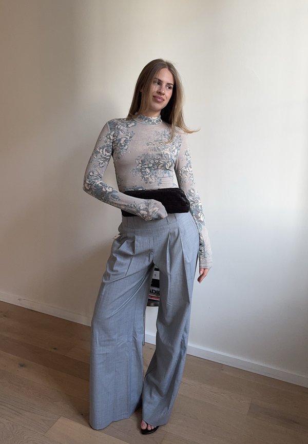 Femeie purtând un top cu mâneci lungi cu imprimeu floral, pantaloni gri cu talie înaltă și croială largă, și sandale negre, stând într-o cameră mobilată minimalist.