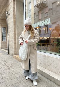Kremowy, oversize'owy płaszcz, szare szerokie dżinsy z postrzępionymi mankietami, białe czółenka za kostkę w szpic oraz fakturowana biała torebka.