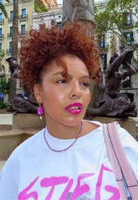 Camiseta blanca con texto rosa, collar de cuentas moradas, pendientes rosas, labios brillantes y cabello rizado castaño. El fondo presenta una fuente de piedra.
